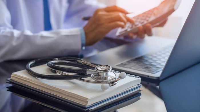 A close-up view of a doctor's hands typing on calculator in front of a laptop. A stethoscope rests on a notebook in the foreground, indicating a medical professional working on patient records or research.