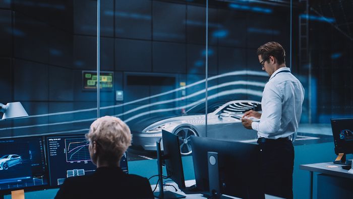 Two people in a high-tech automotive design studio. One stands examining a holographic car projection, while another sits at a computer workstation with multiple screens displaying technical data and designs.