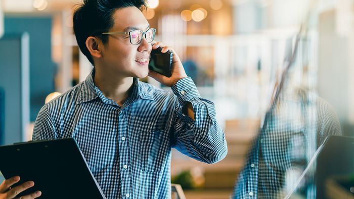 A young business person wearing glasses and a checkered shirt, smiling while talking on a mobile phone. They are holding a tablet or clipboard and standing in a modern office space with blurred background.